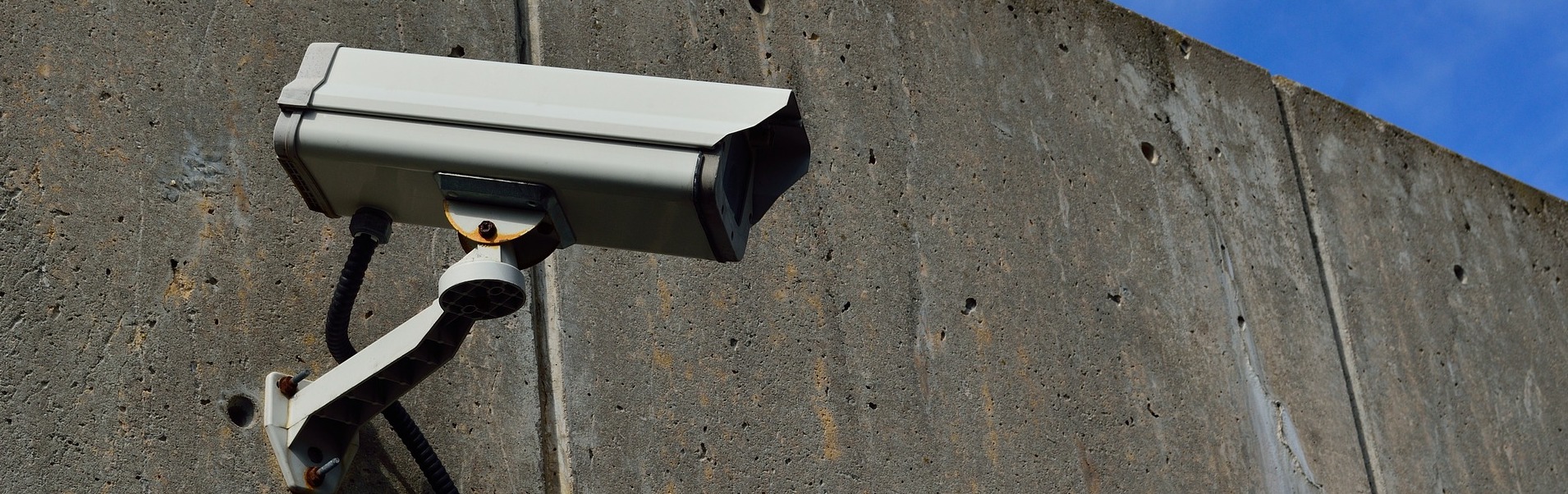 A security camera mounted on a grey, concrete wall with blue sky visible in the background. The camera is positioned to monitor activity, and the wall has visible joints and some small holes. In case of incidents requiring personal injury attorneys, this footage could be crucial evidence.