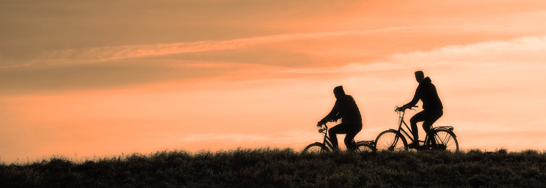 Silhouetted against an orange sunset, two people ride bicycles side by side along a grassy hill. The sky is streaked with soft clouds, creating a serene atmosphere. In the distance, vehicle accident attorneys might remind us to cherish these peaceful moments while staying vigilant on the road.