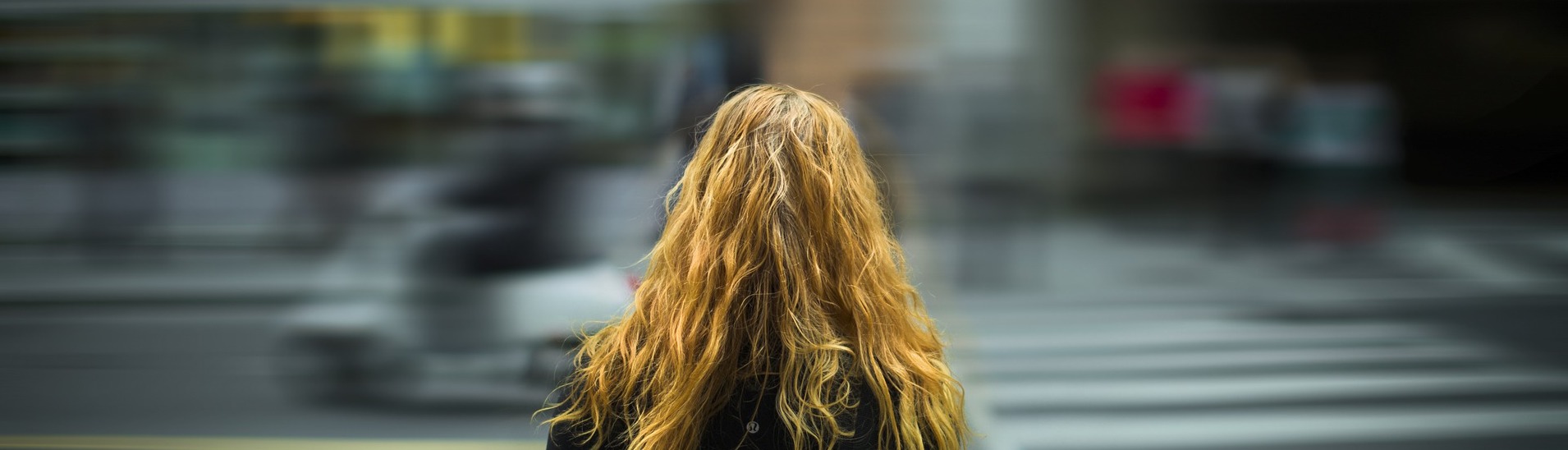 A person with long, wavy blonde hair stands facing away from the camera in an urban setting, gazing at a slightly blurred street with people and vehicles in motion, capturing the busy atmosphere of the city—a scene that might catch the attention of vehicle accident attorneys.