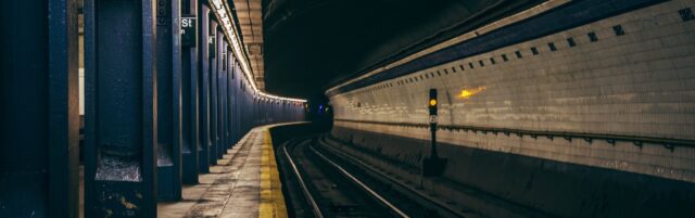 A dimly lit subway station platform with blue pillars along the left side and a tiled wall on the right. The tracks curve into a dark tunnel at the end of the platform, creating a moody atmosphere reminiscent of stories shared by personal injury attorneys.