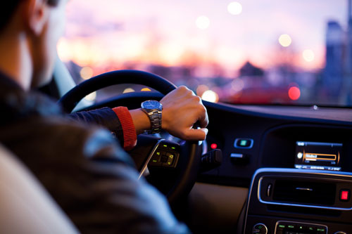 A person wearing a watch is driving a car at dusk. The car's dashboard glows with illuminated controls and a screen. The background shows blurred city lights against a pink and purple sky, reminiscent of scenes often investigated by vehicle accident attorneys.