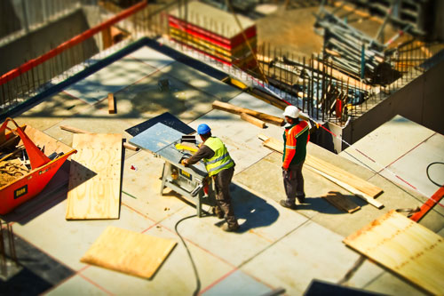 Two construction workers wearing safety vests and helmets work on a construction site. One operates a saw, cutting wooden planks, while the other stands nearby. Various construction materials and tools are scattered around on the concrete floor, ensuring that personal injury attorneys' services are in demand.