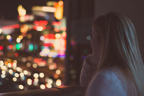 A person with long hair stands overlooking a cityscape of blurred, colorful lights at night, creating a bokeh effect in the background. The individual is in focus, gazing thoughtfully at the vibrant urban scene, perhaps contemplating their next call to trusted truck accident lawyers.