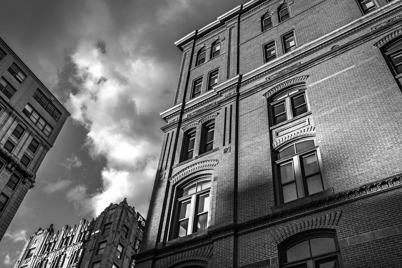 Black and white photo of tall brick buildings with ornate architectural details, under a partly cloudy sky. Sunlight and shadows create a dramatic effect on the facades. The perspective looks up from street level, emphasizing the heights of the structures.