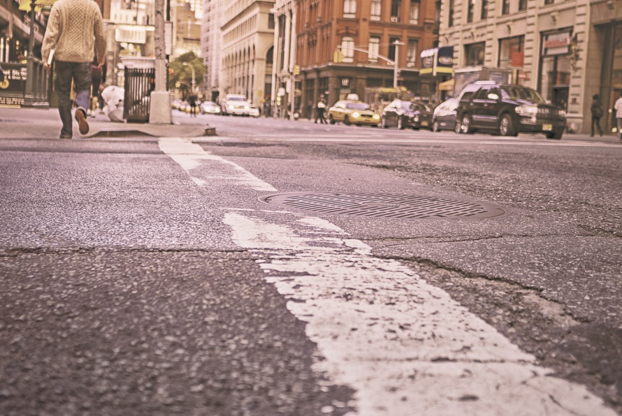 A city street with a focus on the road, showing a white line and a manhole cover. In the background, a person walks on the sidewalk, and several cars are driving by buildings. The scene has a vintage filter.