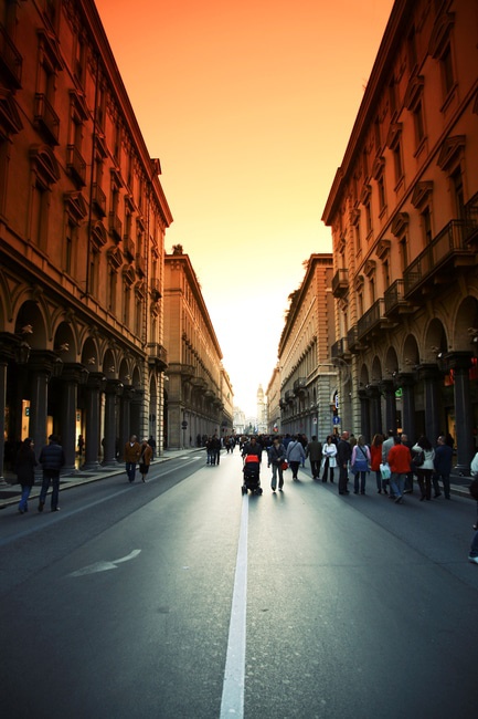 A city street at sunset, framed by tall buildings on both sides, with an orange-yellow sky. People walk down the street, and a person with a stroller is in the center. The architecture features arches and balconies, giving a historic feel.