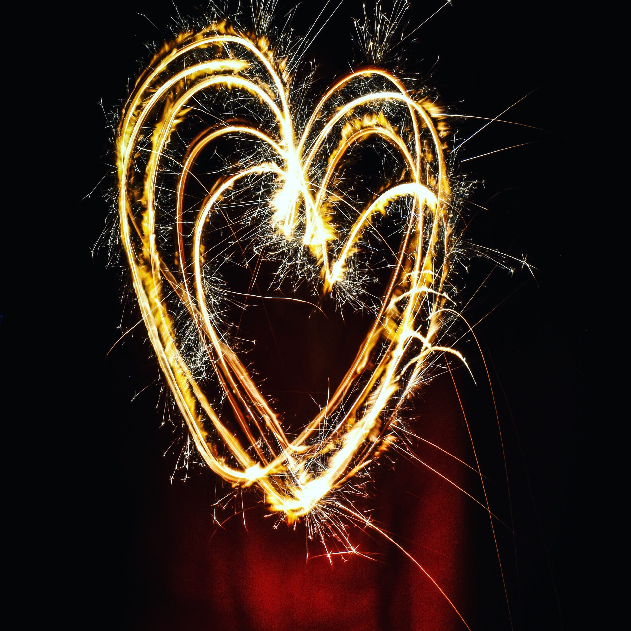 A heart shape is drawn using bright, glowing sparklers against a dark background. The light trails are vibrant and dynamic, creating a warm and lively atmosphere.