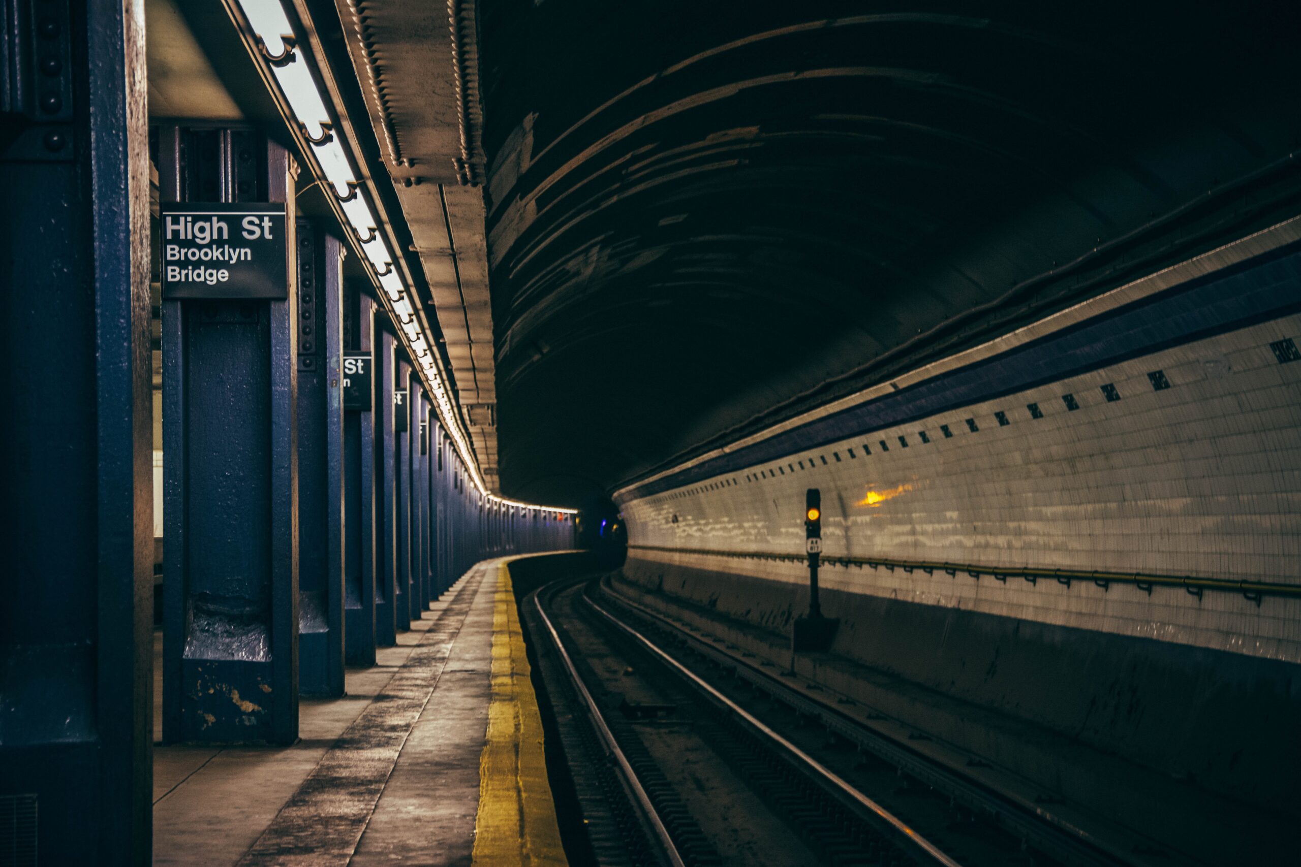 A dimly lit subway station platform of High Street Brooklyn Bridge. The platform is empty, with a curved track disappearing into the tunnel. Blue columns line the left, and a yellow safety line runs along the edge of the platform.