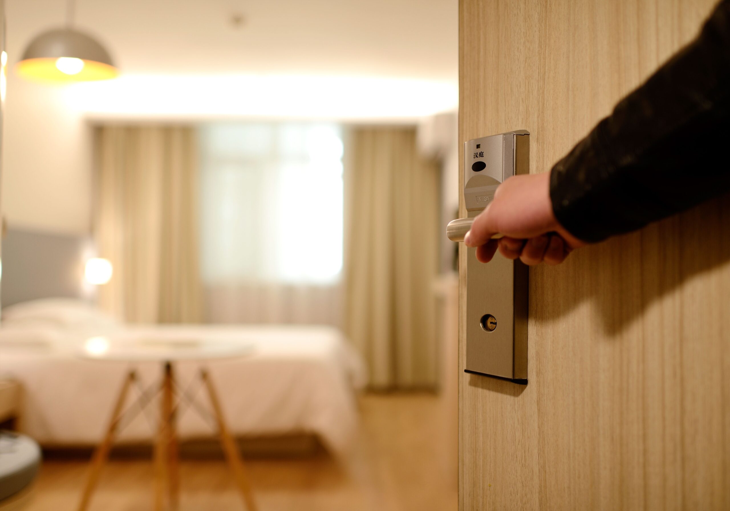 A hand opening a wooden hotel room door with a keycard lock. Inside, there’s a neatly made bed, a desk with a chair, and lightly colored curtains partially covering a window. Warm lighting illuminates the room, creating a cozy and inviting atmosphere.