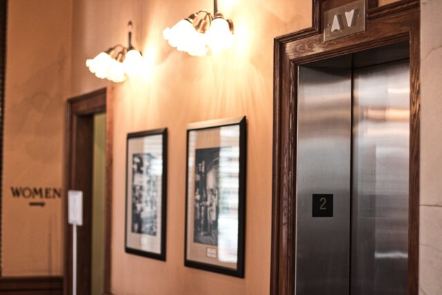 A hallway features a women's restroom door on the left, two framed black-and-white photographs in the center, and an elevator with the sign 