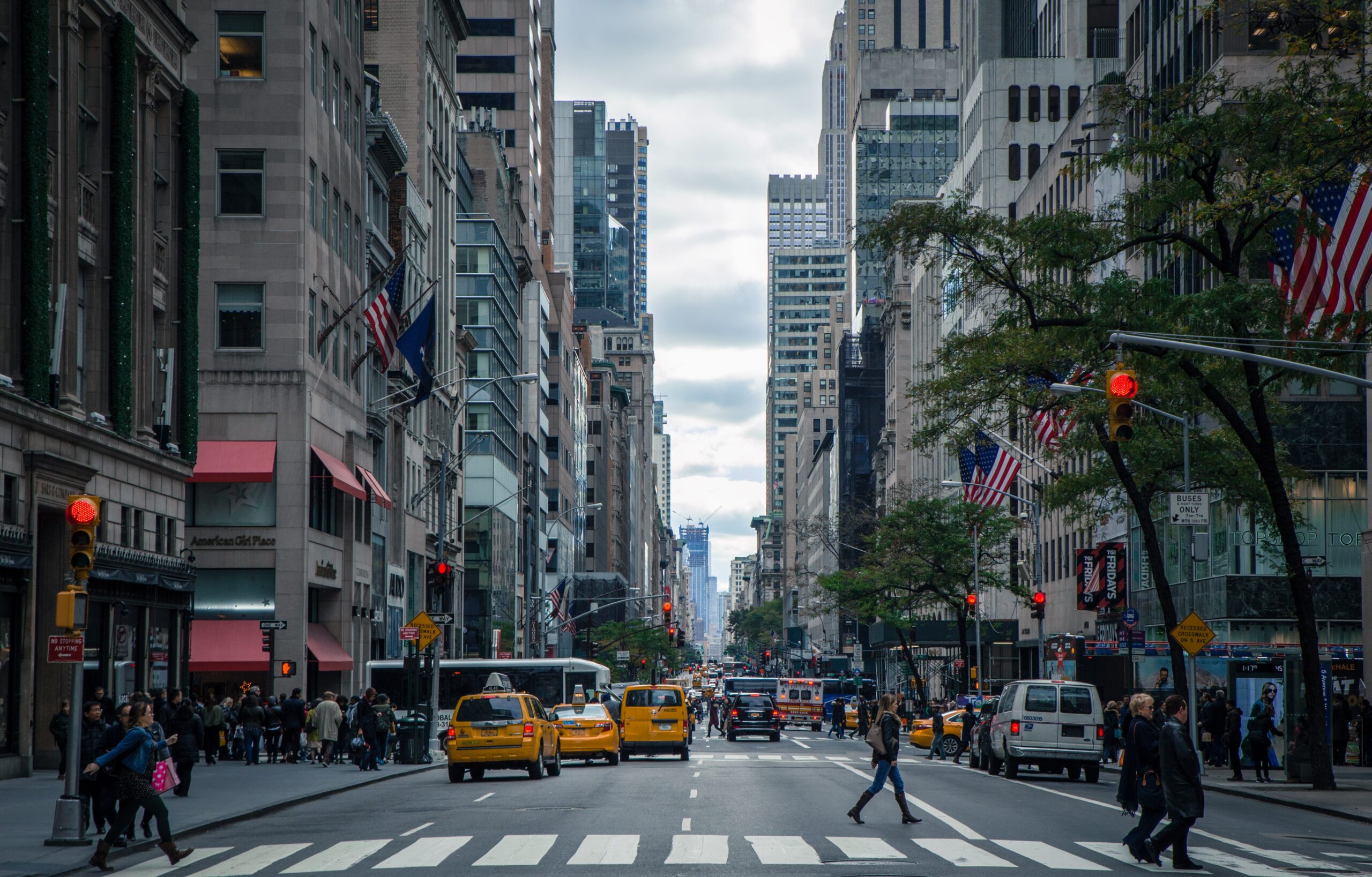 A bustling city street in New York, featuring tall buildings, pedestrians, yellow taxis, and American flags. The sky is overcast and traffic lights are red, with vehicles and people crossing the zebra-pattered intersection.