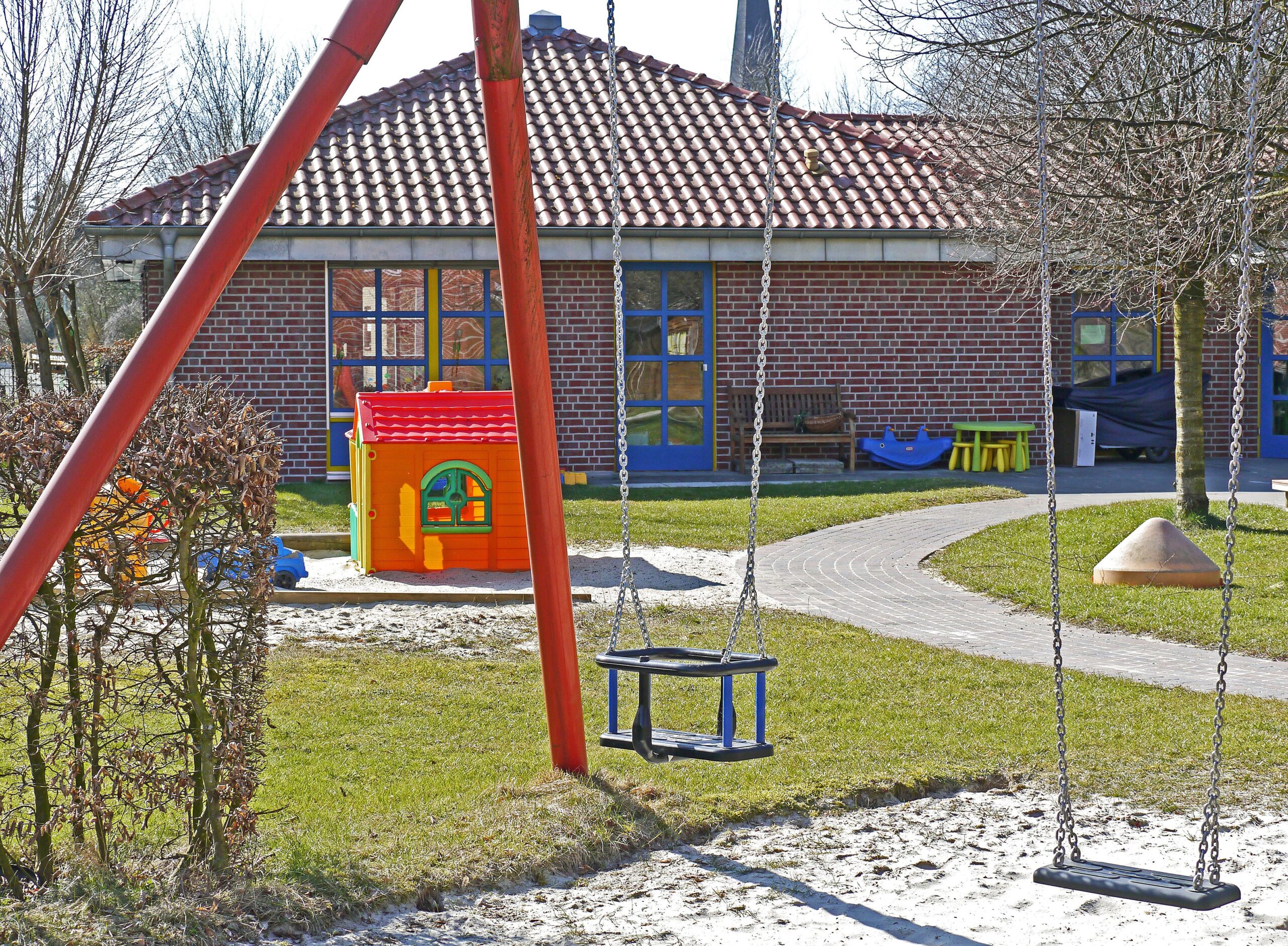 A playground with two empty swings in the foreground. Behind the swings, there is a small, colorful playhouse, and a red brick building with a tiled roof. Trees without leaves surround the area, and a paved path winds through the grass.