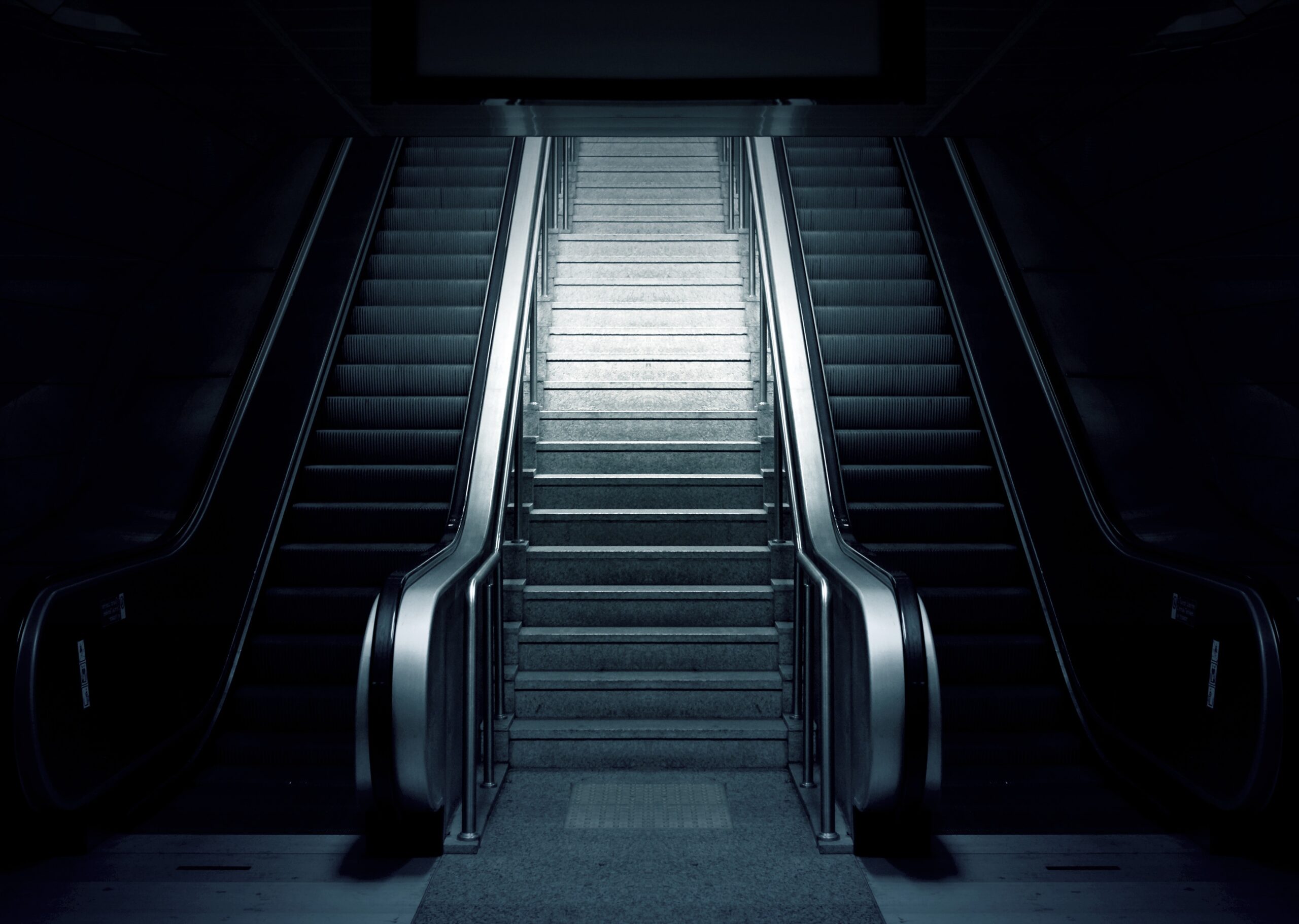 A dimly lit indoor space shows a symmetrical view of two escalators flanking a central staircase. The escalators are not in motion, and the scene has a moody, almost futuristic feel with a strong emphasis on shadows and light.