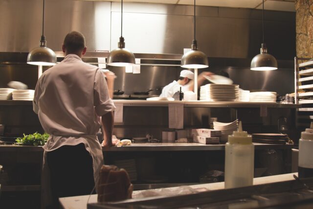 A busy restaurant kitchen with cooks wearing white uniforms and hats preparing food. One cook stands in the foreground while others work in the background, handling plates and ingredients under hanging lights. The kitchen has stainless steel surfaces.