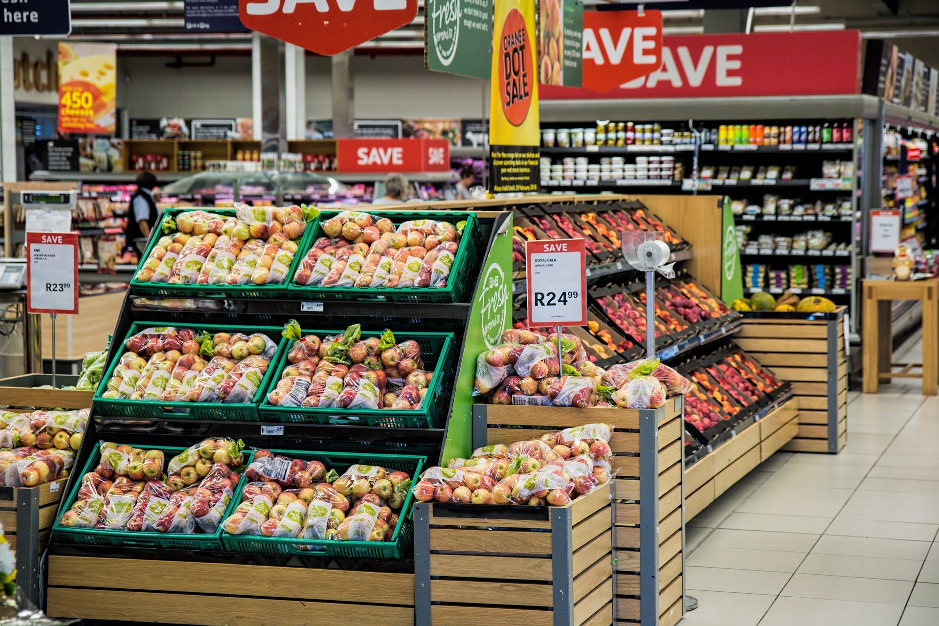 A supermarket produce section with multiple displays of prepackaged fruits, mainly apples, marked with price tags. Shelves in the background have various canned and packaged goods. Red 