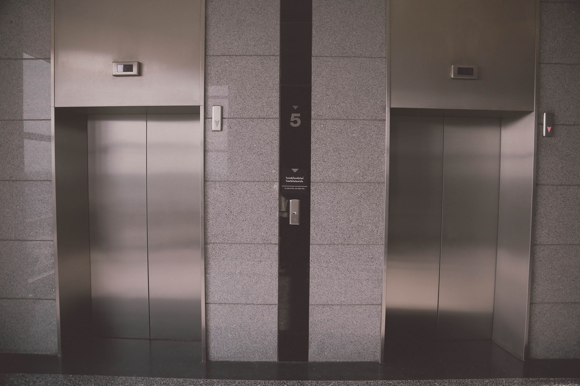 The image shows two closed stainless steel elevator doors located in a modern building lobby. The wall around the elevators is covered in a gray stone finish. A control panel with an indicator shows the number 5 between the two elevators.
