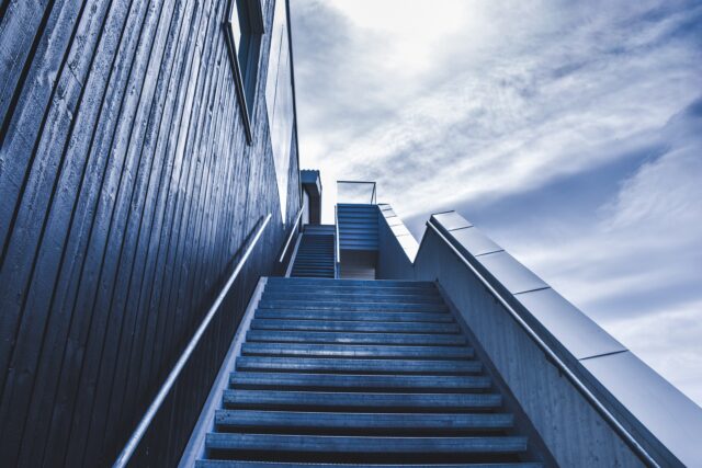 An exterior staircase with metal railings leads up between two modern buildings on a cloudy day. The wooden and metal cladding of the buildings, combined with the overcast sky, highlight the clean lines and minimalist architecture of the scene.