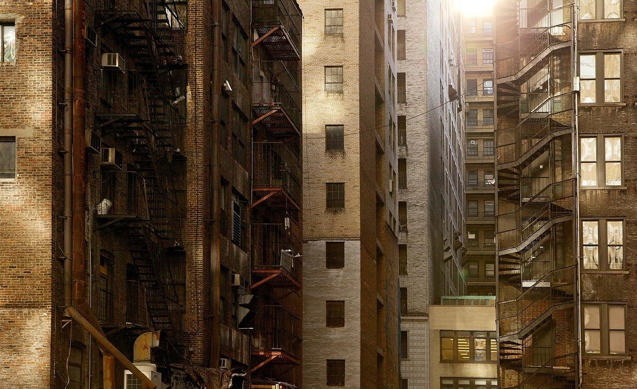 Sunlight peeks through tall, narrow alleyways between old brick buildings with fire escapes and air conditioning units. The scene captures an urban, densely-packed residential area with weathered exteriors and minimal greenery.