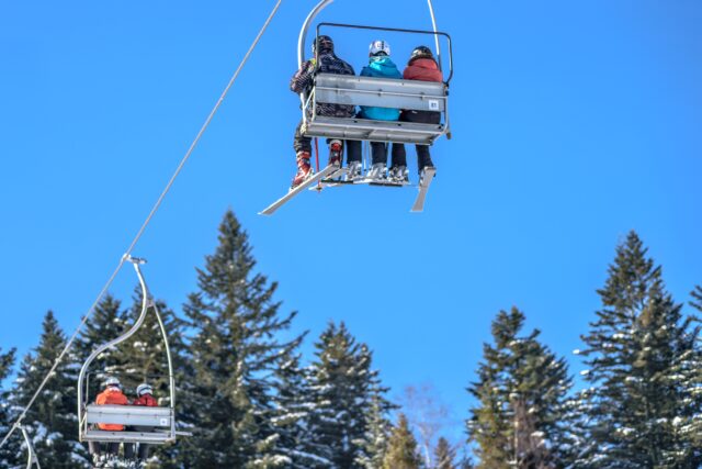 Three people sit on a ski lift chair, ascending a mountain under a bright blue sky. The riders are geared in winter clothing and helmets. Pine trees laden with snow frame the background, with another ski lift chair visible in the distance.