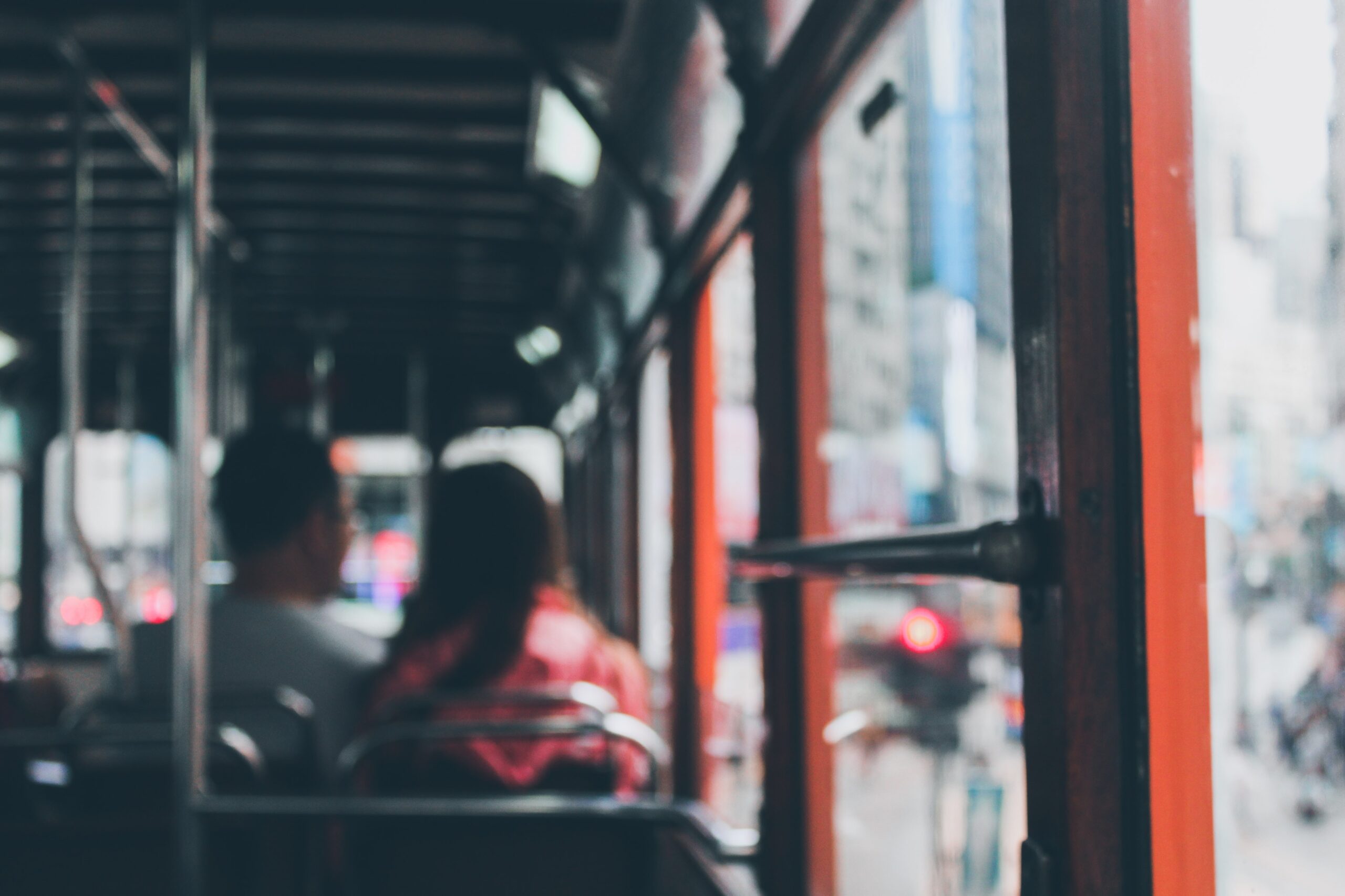 A photo taken inside a bus, slightly out of focus, showing two seated passengers looking toward the window. The bus interior features red poles and metal seats. Outside the window, a busy, urban city scene with blurred buildings and lights is visible.