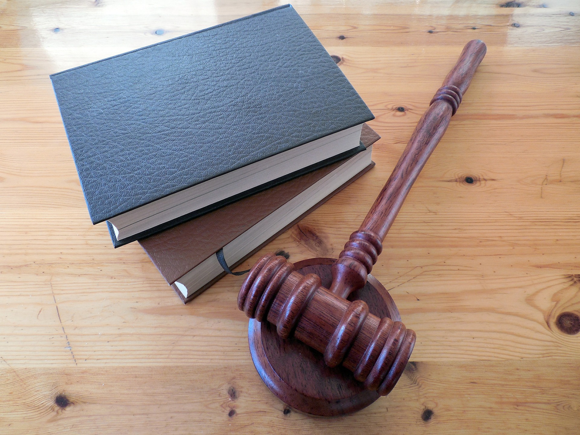 A wooden gavel rests on its sound block on a polished wooden table, next to three stacked books with leather-like covers. The scene suggests themes of law, justice, and legal proceedings.