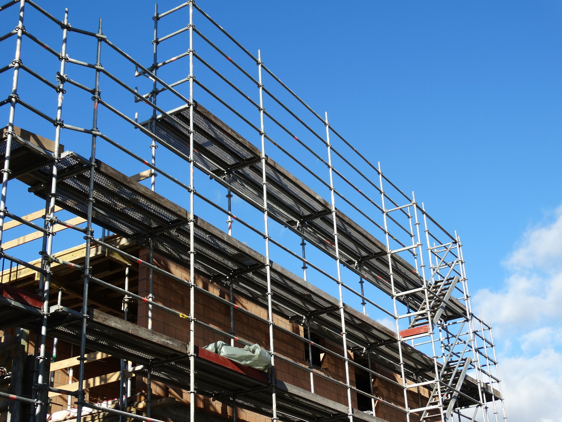 A construction site with metal scaffolding surrounding a partially built structure, set against a clear blue sky. The scaffolding features several platforms at different levels, indicating ongoing building work. Some areas of the structure appear to be brickwork.