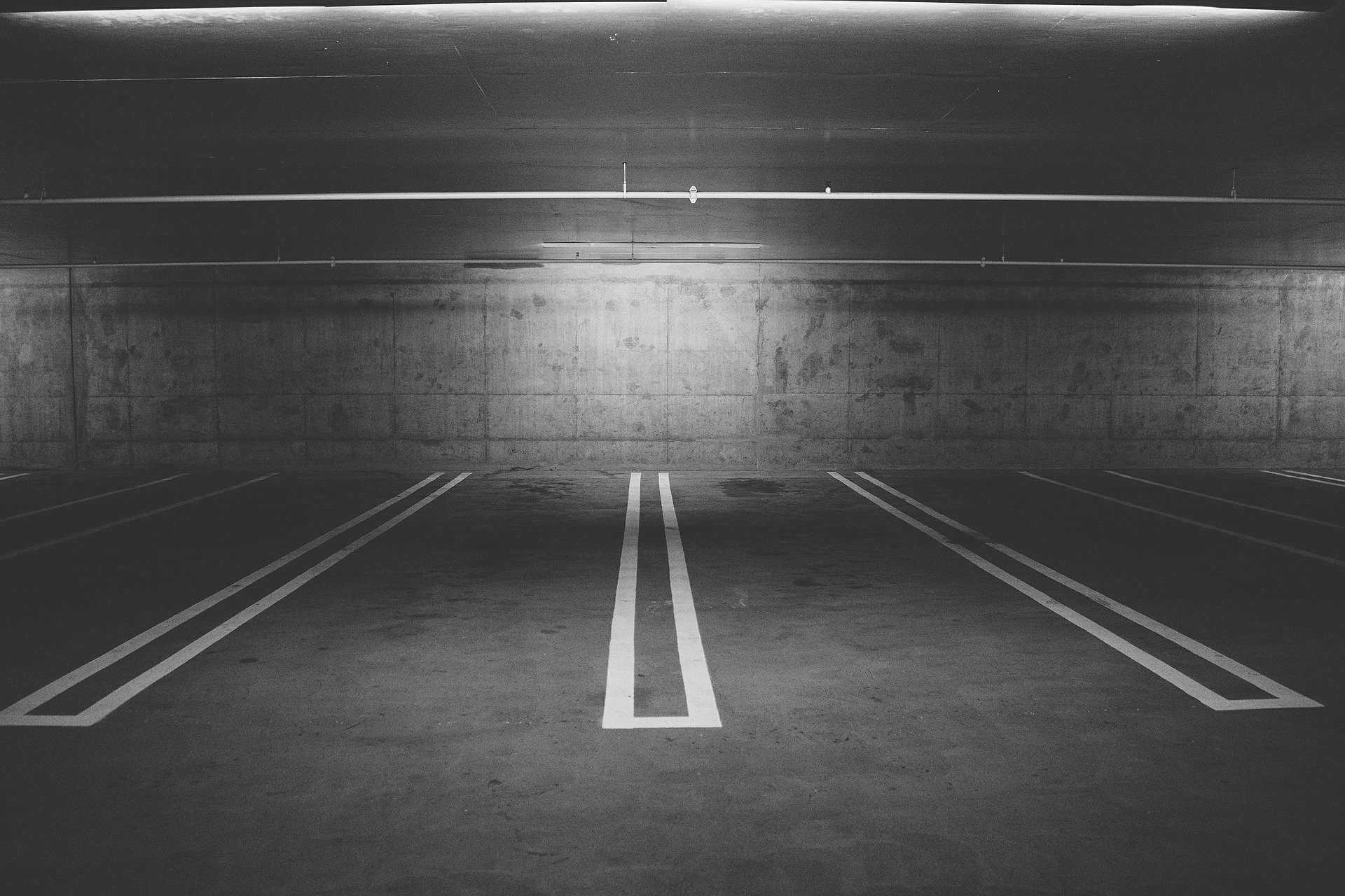 A black and white photo of an empty underground parking garage. The concrete walls and ceiling are bare, and the floor has clearly marked parking spaces with white lines. The lighting is dim, creating a moody atmosphere.