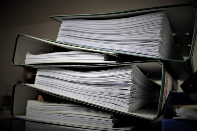 A stack of green ring binders filled with numerous white sheets of paper, piled haphazardly on top of each other. The binders are large and appear to be bursting with documents, indicating an extensive amount of information stored within.