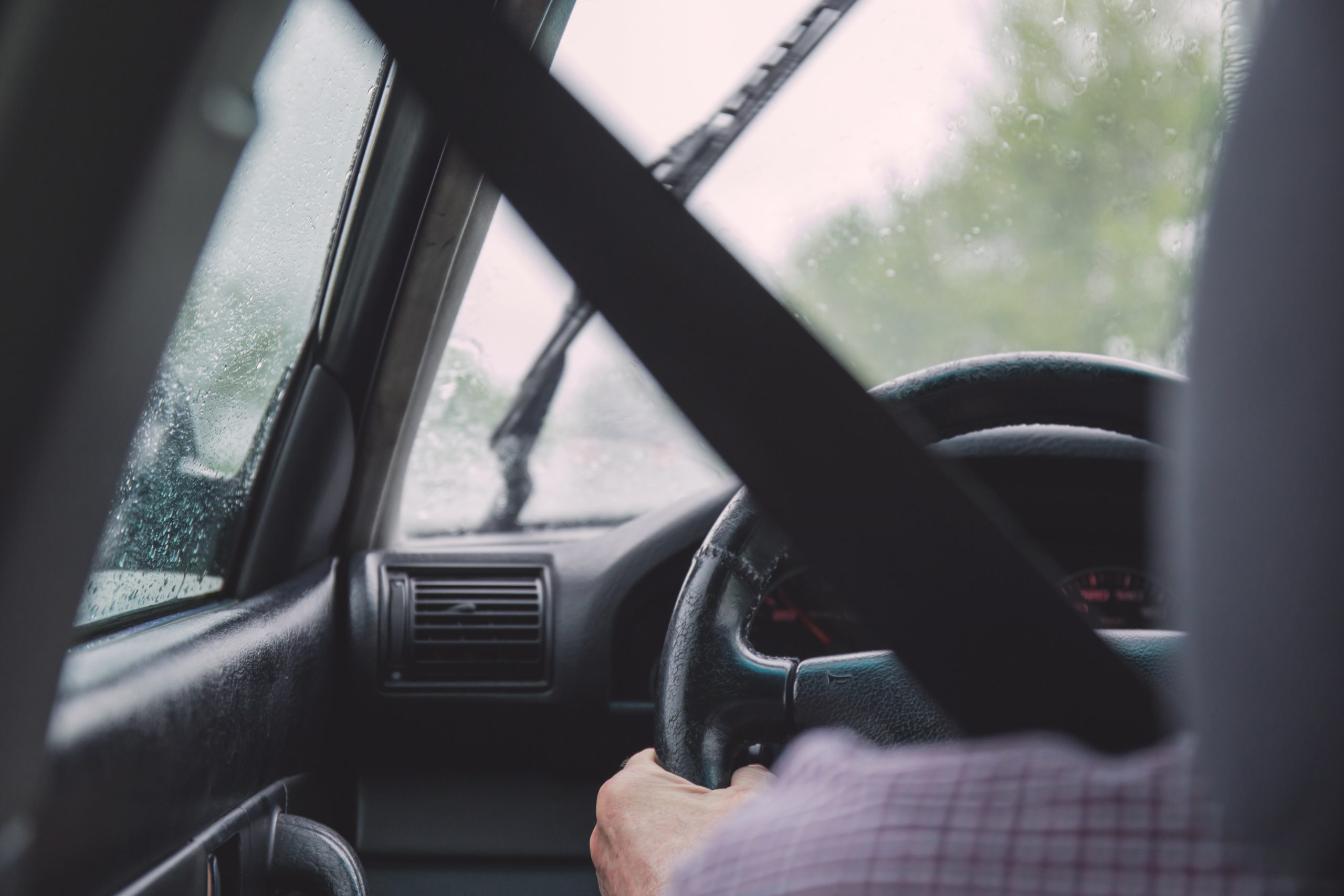 Driver's hands on the steering wheel of a car, with a seatbelt fastened across their chest. The car's windshield wipers are in motion, clearing rainwater from the windshield on a wet day. The view is from the inside of the car, focusing on the driver and windshield.