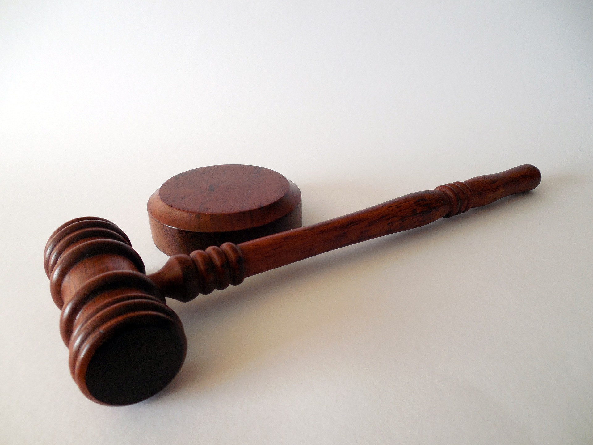 A wooden judge's gavel and sound block rest on a white surface. The gavel, with its handle extending to the right, is positioned parallel to the round, flat sound block, symbolizing law, order, and justice.