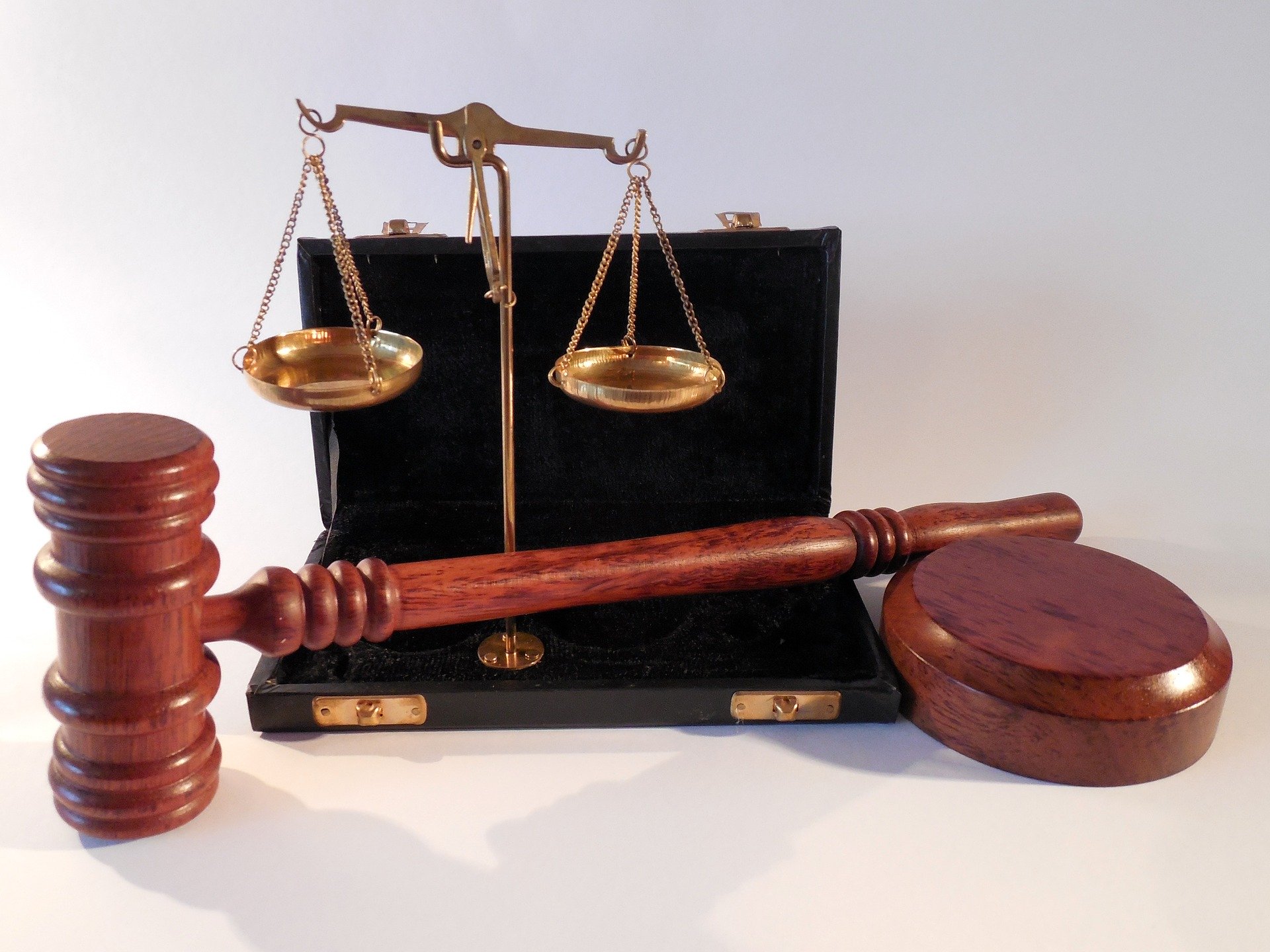 A wooden gavel and sound block are placed in front of a small brass balance scale, all set against a black briefcase. The gavel and balance scale are symbols of justice and law.