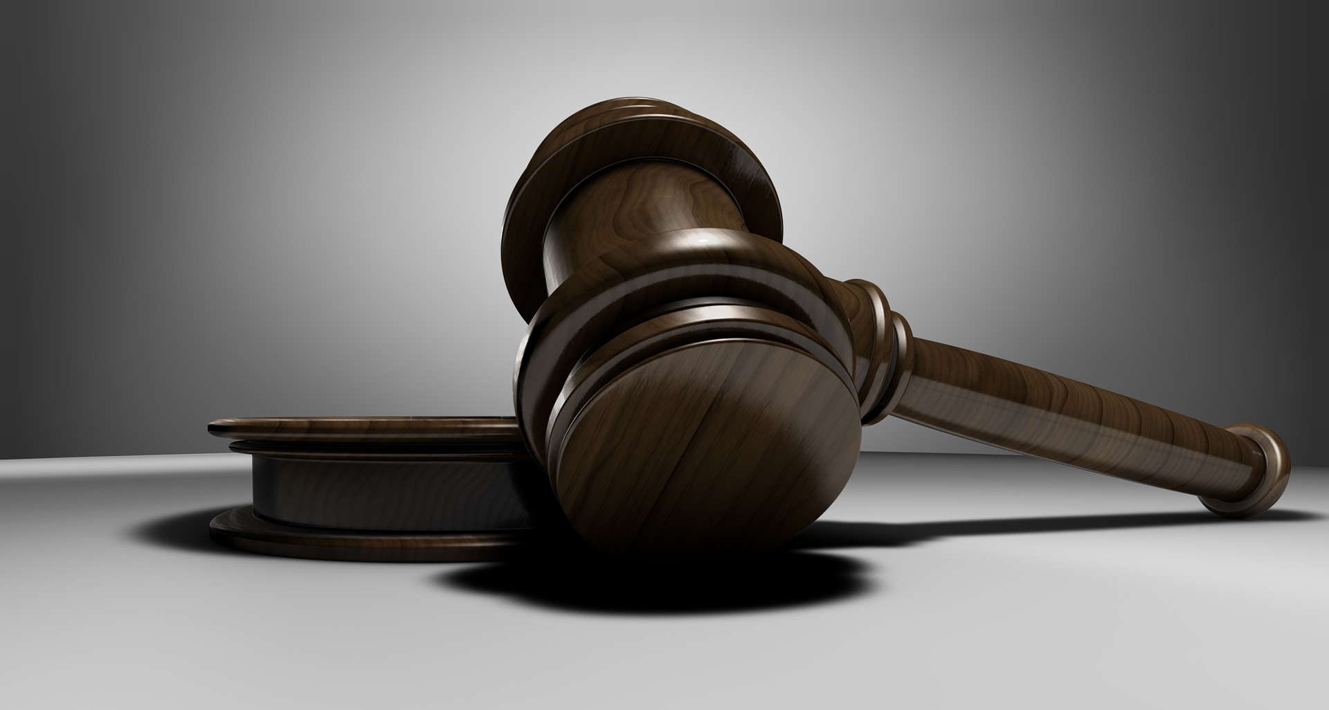 A close-up image of a wooden judge's gavel lying flat on a matching circular sound block against a gradient grey background. The gavel is shown from a low angle, emphasizing its polished surface and detailed grain.