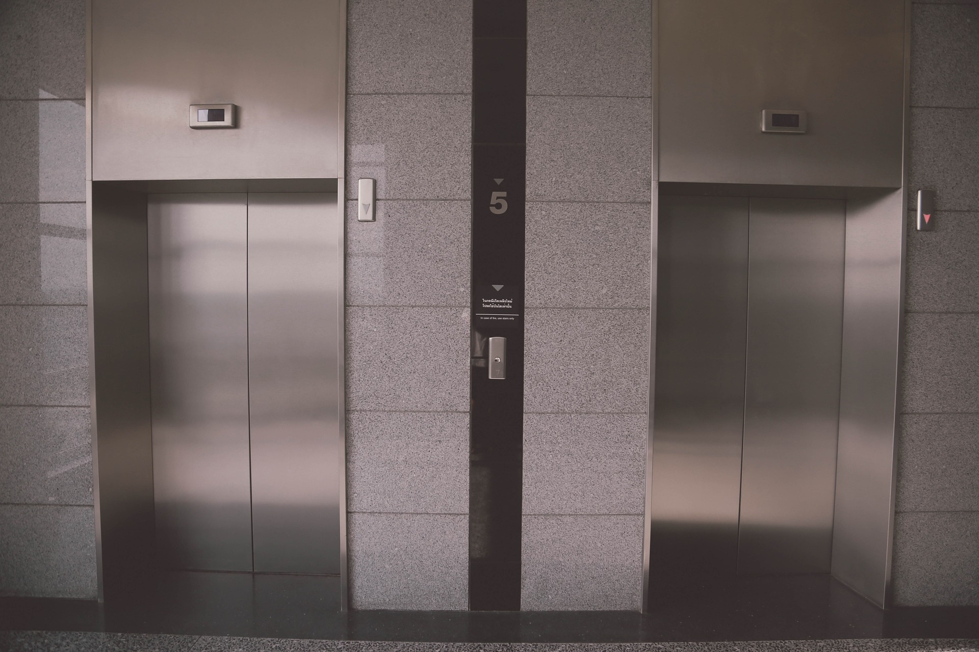 An image showing two closed elevator doors in a modern building lobby. The wall between the elevators is marked with the number 