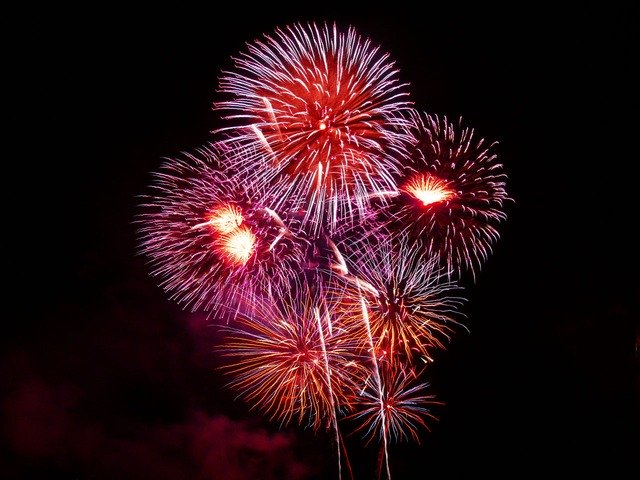 A vibrant fireworks display illuminates the night sky with bursts of red, pink, and purple colors, creating a dazzling and festive atmosphere. The fireworks are captured mid-explosion, forming intricate patterns against the dark backdrop.