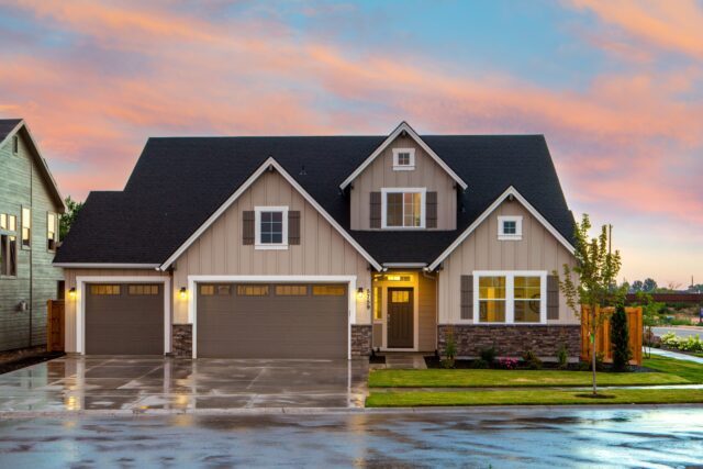 A modern, two-story house with a gray and beige exterior, complete with a dark roof and white trim. The home has two garage doors, a front lawn with a small tree, and neatly manicured landscaping. The sky is vivid with a pink and orange sunset.