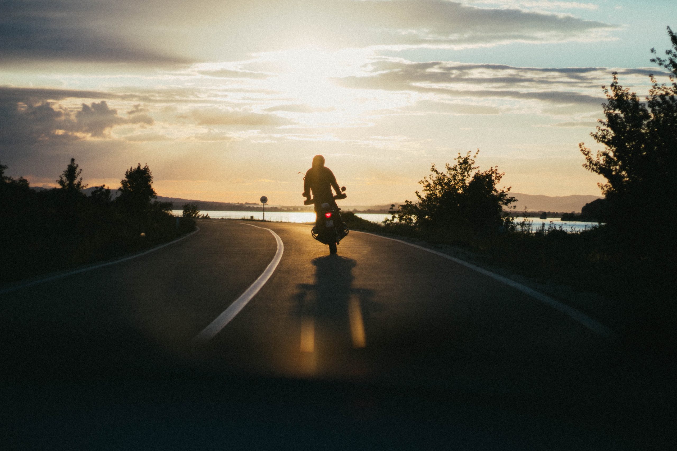 A person rides a motorcycle down a winding road at sunset, casting a long shadow. The road is flanked by trees and bushes, and the sun is low in the sky, emitting a warm, golden light that illuminates the scene. A body of water is visible to the right.