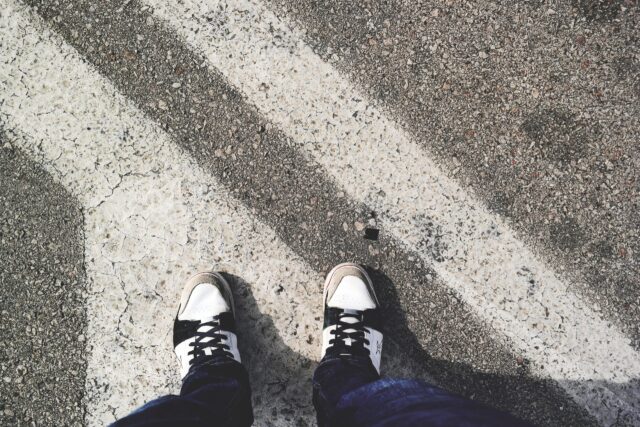 A person wearing white and black sneakers stands on a textured pavement, partially covered by white painted lines. The ground is a mix of gravel and rough concrete, with shadows adding depth to the scene. The person is dressed in dark jeans.