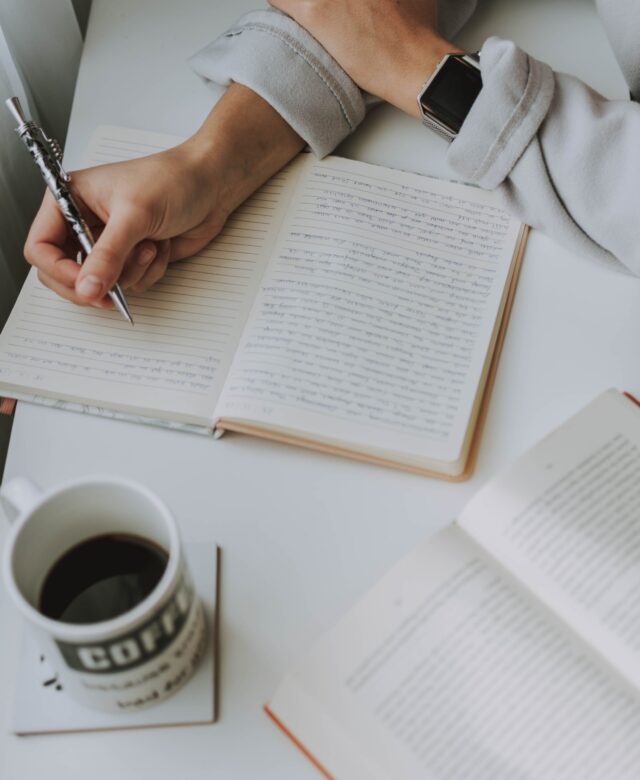 A person is writing in an open notebook placed on a white desk, with a wristwatch on their left wrist. There is a mug labeled 