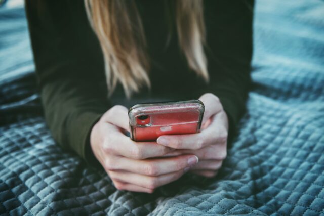 A person with long hair is holding a red smartphone with both hands. The individual is wearing a dark long-sleeved shirt and is seated on a quilted, textured gray blanket. The phone has the Apple logo visible on its back.