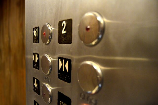 Close-up view of an elevator control panel with illuminated buttons for different floors. The number 2 is prominently visible, and there are buttons with symbols for opening and closing the doors. The panel is metallic with a slightly reflective surface.