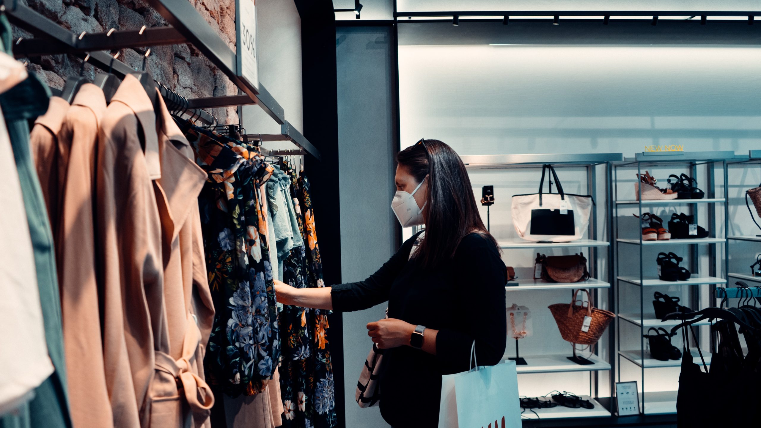 A woman wearing a face mask shops for clothes in a store, browsing through a rack of garments. She is holding a white shopping bag and surrounded by displays of accessories and clothing items against a backdrop of shelves and a brick wall.