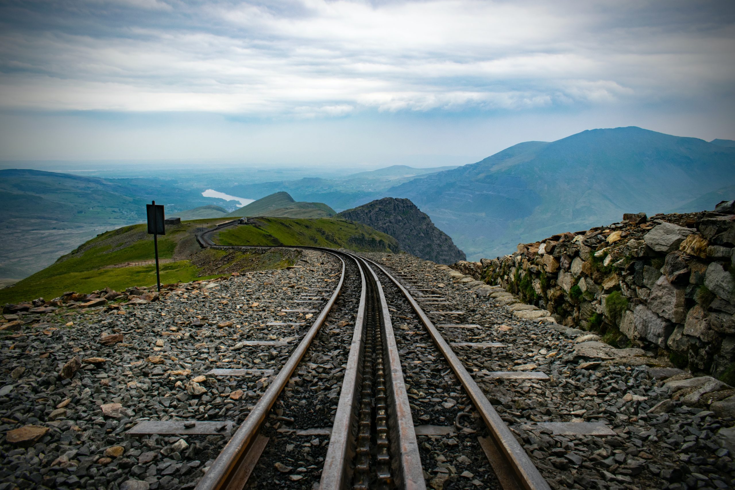 A scenic mountain railway track stretches into the distance under a cloudy sky. Surrounding the tracks are rocky terrain and lush green slopes, with distant rolling hills and a lake visible in the background. A stone wall runs parallel to the tracks on the right.