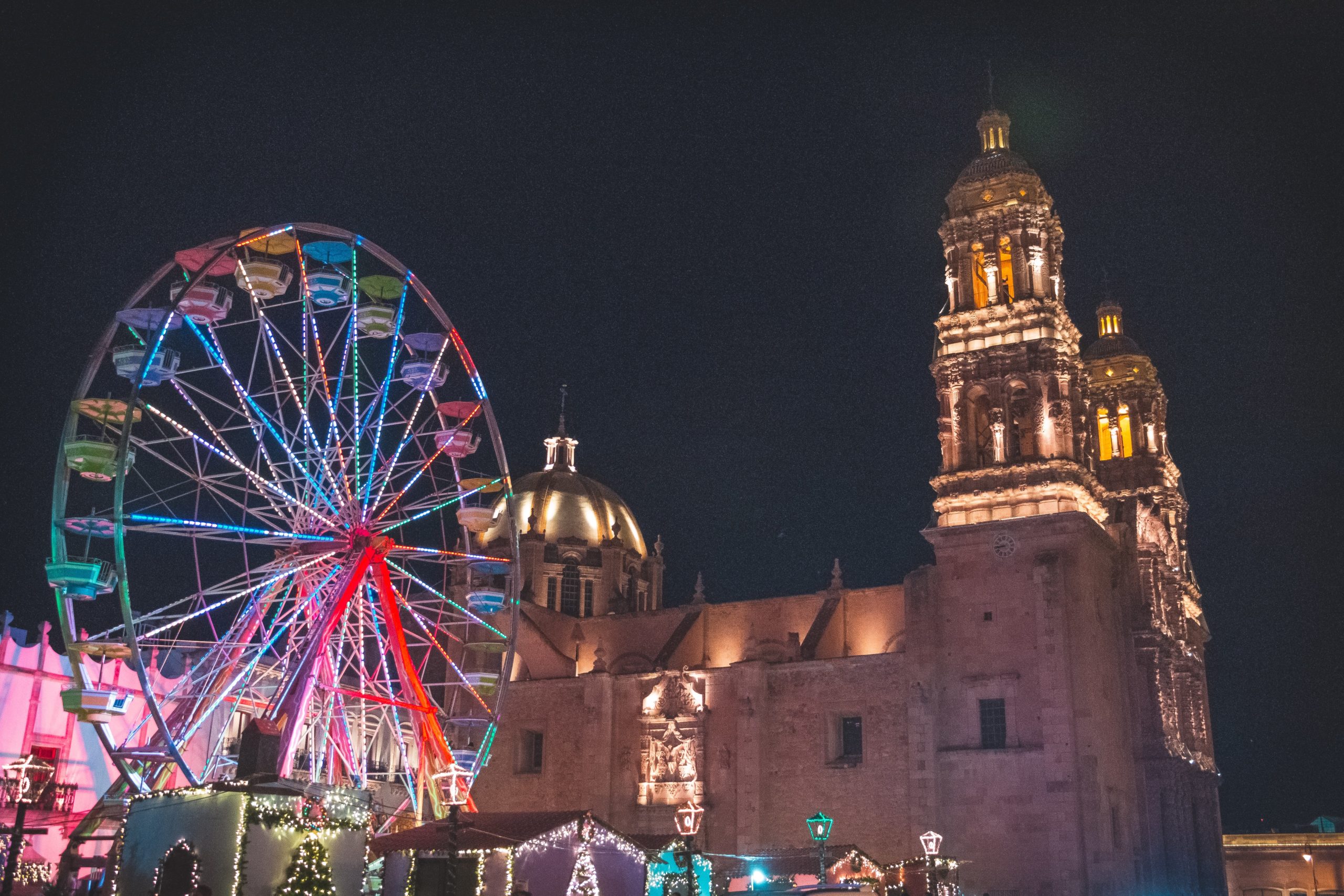 A night scene featuring a brightly lit Ferris wheel with multicolored lights to the left and an ornate, illuminated cathedral with twin towers to the right. The structures are surrounded by festive decorations and illuminated arches.