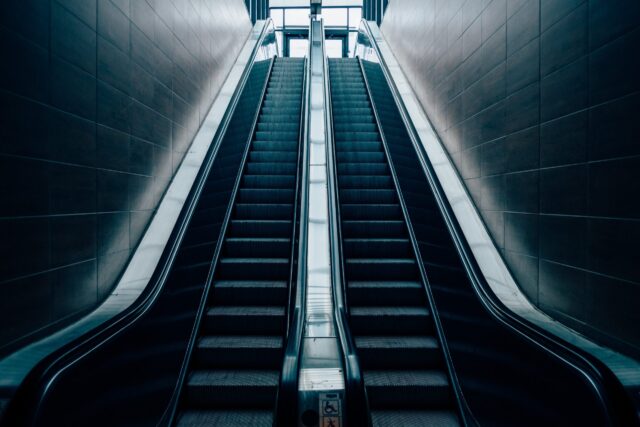 A modern, empty escalator and a flight of stairs parallel to each other ascend towards a bright, illuminated area at the top. The surroundings have a sleek, minimalist design with smooth walls and stainless steel handrails.