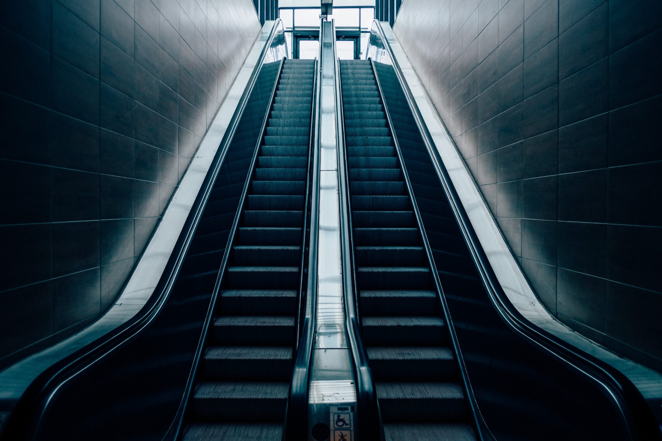 A modern, empty escalator and a flight of stairs parallel to each other ascend towards a bright, illuminated area at the top. The surroundings have a sleek, minimalist design with smooth walls and stainless steel handrails.