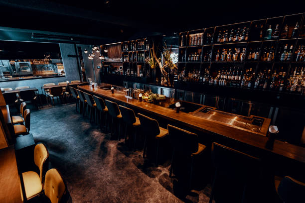 A modern, dimly-lit bar with a long wooden counter lined with high-backed chairs. Shelves stocked with various bottles of alcohol and glassware are visible behind the counter. The bar features a sleek, contemporary design with a cozy, inviting atmosphere.