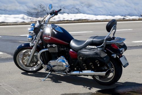 A black and red cruiser-style motorcycle is parked on the side of a road. The motorcycle has a chrome engine and exhaust, a cushioned seat with a backrest, and saddlebags. In the background, there's snow along the roadside.