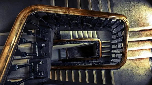 A view looking down a spiral staircase with dark-colored steps and a wooden handrail. The design creates a geometric, repeating pattern. The lighting casts shadows, highlighting the stairs and railings, giving the scene a dramatic and somewhat moody atmosphere.