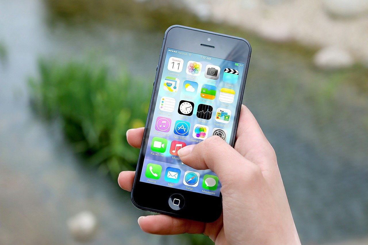 A hand holding a black smartphone with a visible home screen. The screen displays various colorful app icons, including the clock, calendar, photos, camera, weather, and settings apps. The background is outdoors with blurred greenery and water.