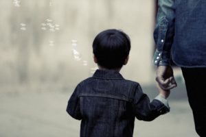 A young child wearing a denim jacket is seen from behind, holding hands with an adult dressed in similar attire. The background is blurred, and a few bubbles are floating around them in the air, creating a serene and tender atmosphere.